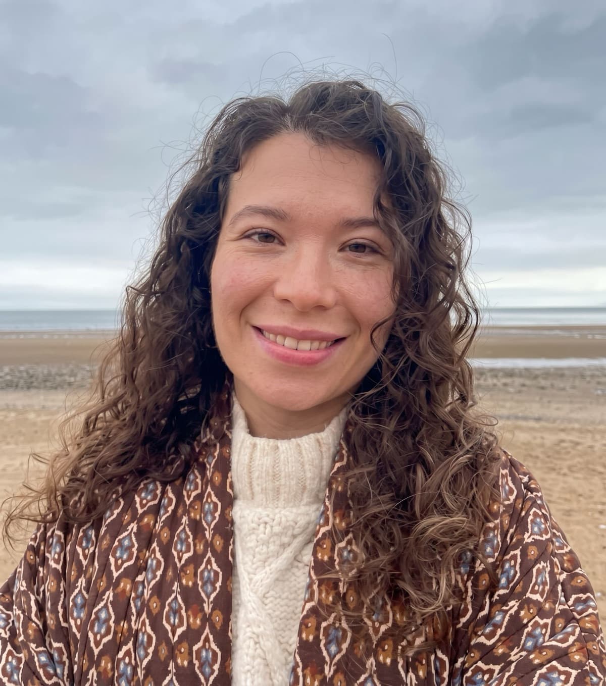 Gabi Machado smiling on a beach in a cream jumper and patterned jacket